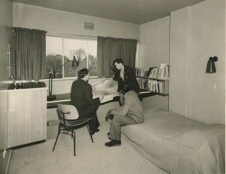 Three students wearing suits in a dorm room chat and read a newspaper.