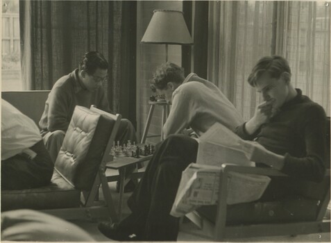 Two young students in the background are playing chess. A young student in the foreground is sitting to the right of frame reading the paper. 