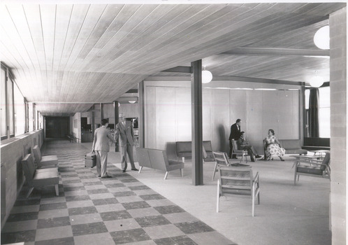 In the background two men in suits shake hands on the left, and two men and a woman sit chatting on the right