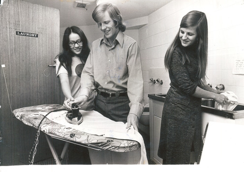 A man irons a shirt whilst a woman to his right assists. A woman washing clothes to their left looks on. 