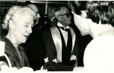 Two women speak in the foreground, framing a man in formal dress in the centre background.