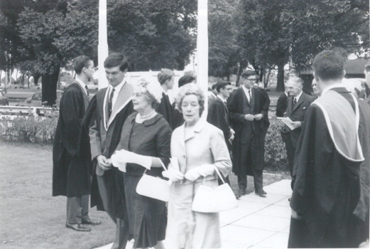 Two elderly women and several men in formal attire accumulated on grass and pavement, with a treed park behind them