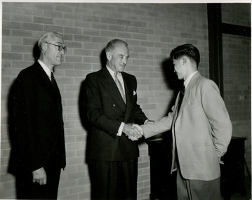 Two men shake hands whilst a third man looks on warmly from the left. All wear suits, and stand before a brick wall.