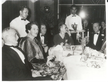 Several people seated around a dining table in formal attire, served by two male waitstaff.