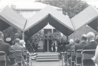 Two columns of guests are seated outdoors, leading up to a marquee, under which a man stands, and another sits in academic attire.