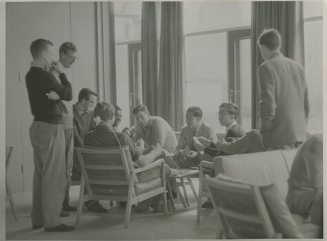 Group of male students gathered around a table. Some are sitting and some are standing. They're all looking at one sitting student holding an unidentifiable object the size of a deck of cards.