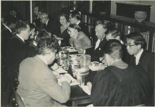 A mix of 15 students, older gentlemen and ladies, sitting around a table eating and chatting. The students are wearing graduate robes. The men are wearing suits. One lady is in a fur coat. 