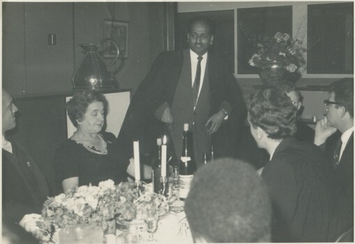 A well dressed Singaporean man standing in and speaking to a table of other well-dressed people. 