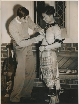 One male student assists another male student put on a costume. The costume appears to Thai inspired. 