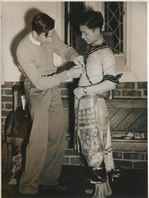 One male student assists another male student put on a costume. The costume appears to Thai inspired. 