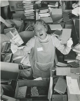 A photograph from above the subject. An exasperated older woman in an apron and white gloves holding up two books is standing in a sea of boxes of books. 