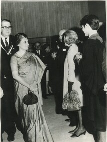 Prime Minister of India Indira Gandhi wearing a sari and holding a small evening bag. A very tall Warden of International House Sam Dimmick  standings a suit behind her. She looks upon a line of well-dressed students eager to greet her. 
