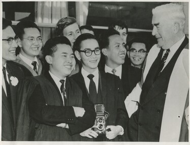 A group of younger male students in suits, one with a camera, standing in front of Prime Minister Robert Menzie, an older man in a suit and oxford robe.