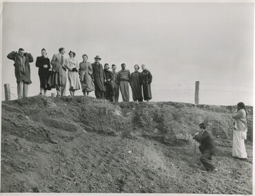 A group of men and women standing in windy weather at the edge of what appears to be a pit. Two people take their photo from below. 