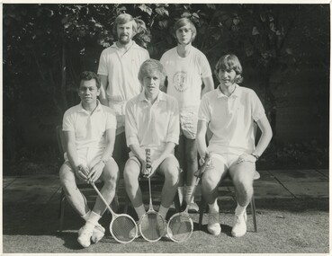 A group of five students. 2 standing , 3 sitting in chairs in front of them. In white collared shirts and shorts. Holding tennis rackets. 