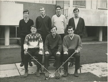 A group of 7 young male students. 4 standing and three sitting in front of them in chairs. The student sitting on the left and the one on the right are both holding golf clubs. All the students are dressed in business casual golf attire. 