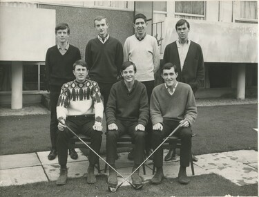 A group of 7 young male students. 4 standing and three sitting in front of them in chairs. The student sitting on the left and the one on the right are both holding golf clubs. All the students are dressed in business casual golf attire. 