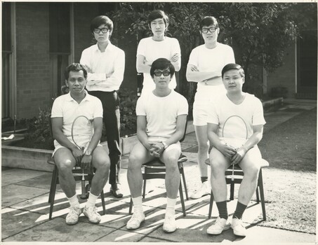 3 young male students standing in white t-shirts behind a row of 3 sitting male students in white t-shirts. Two of the sitting students are holding tennis rackets. 