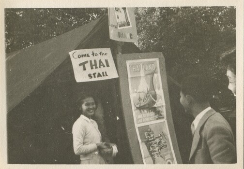 A smart dressed smiling young woman from Thailand stands in front of a tent with posters advertising Thailand. We see the left side-profile of two young males in the right bottom corner. 