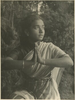 A South Asian woman in a saree holding a traditional dance pose in front of some trees and bushes. 