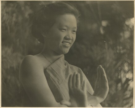 A Pacific Islander female dancer in a dance pose in front of some bushes. 