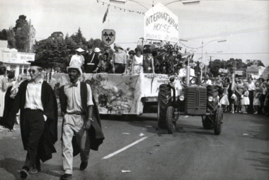 Two men in academic gowns walk on a main street, followed by a tractor pulling a float entitled 'International House' full of people