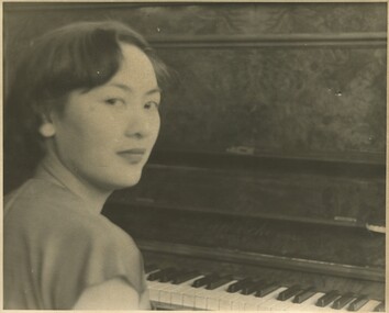 A close-up photo from behind a woman sitting at a piano. She is turning over her right shoulder and looking at the camera. She has a serious and pleasant expression.