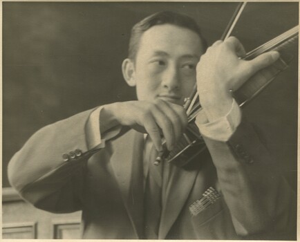 A young Asian man in a suit is pictured delicately holding his violin as if playing or about to play. He looks off to the distance on his left. 