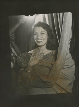 A smiling woman of Polynesian decent covers herself with a velvet theatre curtain, as if hiding her outfit. She looks directly at the camera. Someone is hold a light high above her head.  