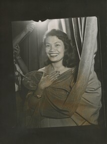A smiling woman of Polynesian decent covers herself with a velvet theatre curtain, as if hiding her outfit. She looks directly at the camera. Someone is hold a light high above her head.  