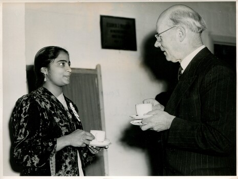 A well-dressed young Sri Lankan woman holding a white mug and saucer speaking to a well-dressed older male, also holding a white mug and saucer. 