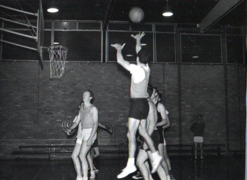 Man shoots a basketball goal in a darkly lit indoor basketball court, as other players watch