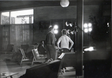 A man and woman face away from the camera at a distance. They are in a large room, surrounded by filming equipment and lights.