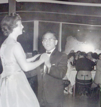 Male student and female in formal attire dance in the foreground. The male reaches the female's shoulder.