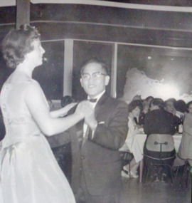 Male student and female in formal attire dance in the foreground. The male reaches the female's shoulder.