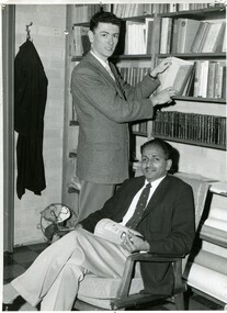 Two well-dressed men, posed candid. One is standing at a book shelf with a book open, looking over his shoulder. The other is sitting with an open magazine. 