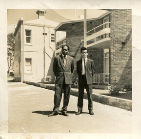 Two men standing outside in suits on a sunny day in front of a brick building. 