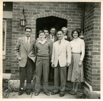 A well dressed group of 5 young men and 2 women standing in front of an entrance to a brick house. 