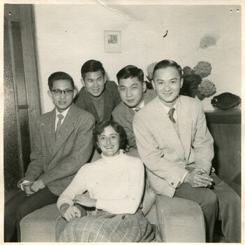 A smiling young woman sits on a sofa chair with 4 men circled around her for the photo. 