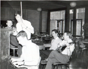 One man plays piano in the foreground, one sings and one plays guitar over his right shoulder, and two sing over his left