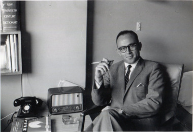 Seated man in a suit and glasses smiles gently at the camera. A desk with a telephone, radio, and book sits behind him.
