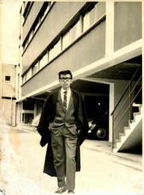Young Asian student in with glasses in a suit and graduation robe standing outdoors in front of a tall building. The photograph has been yellowed from age and slightly tattered.