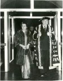 An older woman in a fur coat walking alongside an older man in Oxford graduation robe regalia. 