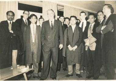 Around fifteen young male students in suits and Oxford robes standing around an older gentleman in a suit. 