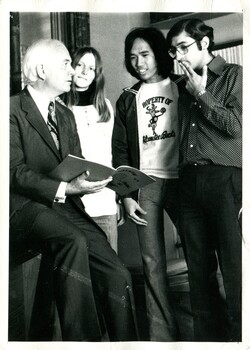 An older man on the left side of the photograph is sitting on a the arm of a chair, he has a magazine open, he is looking at a group of three casually dressed students. One young woman, two young men. 