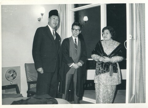 A man in a suit and sangkok (hat), a younger male in suit, Oxford robe, and glasses, a woman in 2-piece evening wear and fur shawl. They are standing by an open window at night. 