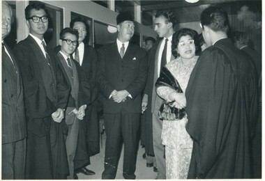 Group of six young men in suits and Oxford robes standing in a semi-circle either chatting to the Sultan, chatting to his wife, or looking at the camera. 