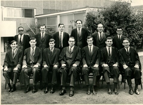 A row of seven men in suits and Oxford robes sitting in front of six standing men in suits and Oxford robes. The group is outside on the International House campus. 