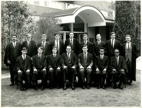 Seven men sitting in chairs in suits and Oxford robes in front of nine standing men in suits and Oxford robes. The group is outside posing for this formal photo. 