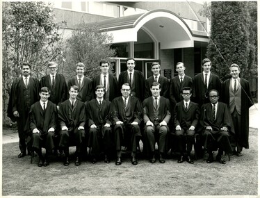 Seven men sitting in chairs in suits and Oxford robes in front of nine standing men in suits and Oxford robes. The group is outside posing for this formal photo. 